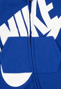 Modrá mikina s kapucňou s bielym logom Nike, predný zips a veľká predná kapsa. Vyrobená z mäkkej látky s posilnenými švami a hladkým povrchom.