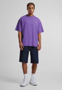T-shirt violet surdimensionné avec broderie dorée, associé à un short en denim foncé et des chaussures de sport blanches. Fond uni, éclairage neutre.