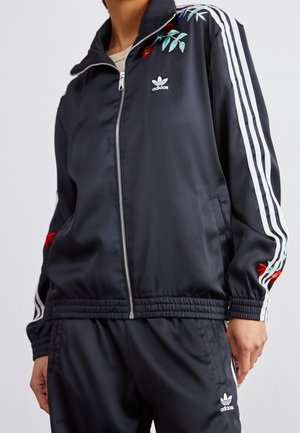 Osoba ubrana w czarny dres Adidas z białymi paskami i kwiatowym haftem na ramionach i rękawach kurtki.