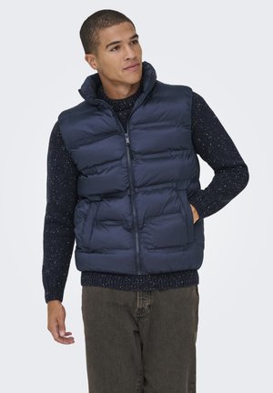 Chaleco acolchado marino con cuello alto, cierre de cremallera y bolsillos laterales. Suéter de punto texturizado debajo, combinado con pantalones caqui.