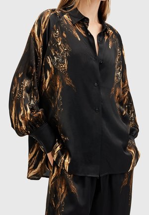 Mujer con camisa negra de satén con botones y pantalones decorados con patrones de plumas y mariposas en bronce, con las manos en los bolsillos, sobre un fondo liso.