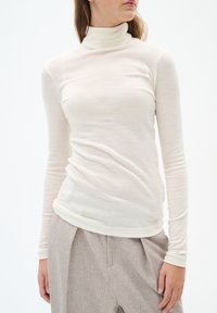 Long-sleeve ivory turtleneck shirt in lightweight, stretchy fabric with a smooth texture, paired with light gray pleated trousers.