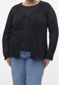 Mustan neulecardiganin avonaisella edellä, monimutkaisella auringonsädekuviolla, pitkillä hihoilla ja narusuljennalla; päällä musta paita ja siniset farkut.