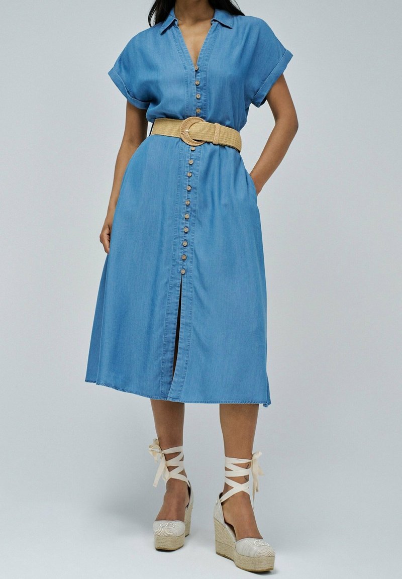 Lichtblauwe denim jurk met korte mouwen, voorste knoopsluiting en een middellange rok. Draag met een brede beige riem en sleehakken.