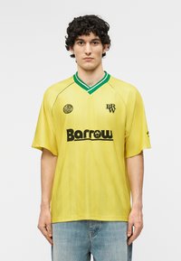 Joven con cabello rizado que lleva una camiseta deportiva amarilla con cuello verde, logos en negro y jeans azul claro sobre un fondo liso.