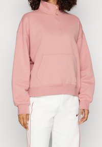 Sudadera de color rosa claro con media cremallera en el cuello, bolsillo tipo canguro y puños acanalados. Tejido suave y texturizado.