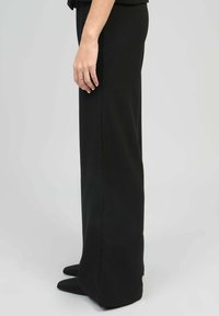 Pantalons noirs à jambes larges en tissu lisse et texturé. Présentent une taille haute et une coupe droite, accompagnés de chaussures noires à bout pointu.