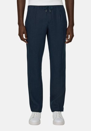 Adulte portant un pantalon à cordon de serrage bleu marine et des baskets blanches, debout devant un fond clair uni.