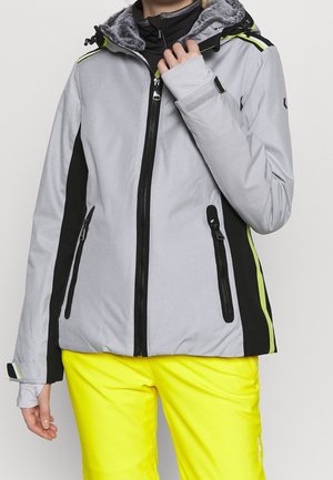 Personne portant une veste d'hiver gris clair et noire avec une capuche bordée de fourrure et un pantalon de neige jaune vif, ajustant la fermeture éclair de la veste.