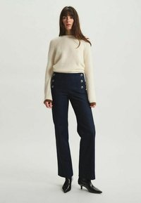 Pantalons bleu marine taille haute avec des boutons à l'avant, en tissu texturé. Associés à un pull à côtes crème et des talons aiguille noirs.