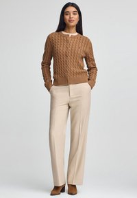 Kvinna klädd i en brun kabelstickad kofta, beige vida byxor och bruna stövlar med klack, stående med händerna i fickorna mot en enkel bakgrund.