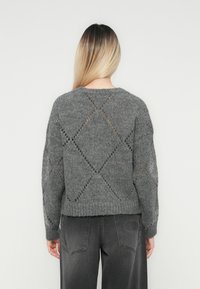 Femeie cu păr drept ombre, purtând un pulover tricotat gri cu găuri în formă de diamant și blugi gri închis, stând în fața unui fundal uniform.