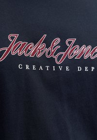 Το λογότυπο Jack & Jones με κόκκινα καλλιγραφικά γράμματα πάνω από το «Creative Dep» με λευκά κεφαλαία γράμματα σε σκούρο υφασμάτινο φόντο.