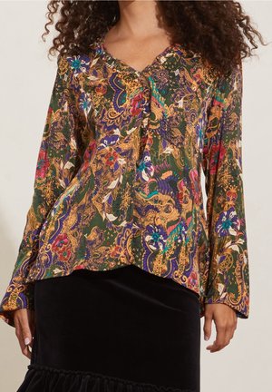 Femme portant un chemisier à manches longues avec un imprimé floral paisley vert, or, violet et rouge, associé à une jupe noire en velours.