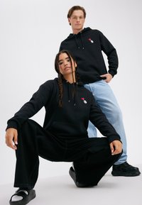 Black hoodies with front pockets, drawstrings, and an embroidered design. Model wears black wide-leg pants and black sneakers, standing on a white background.