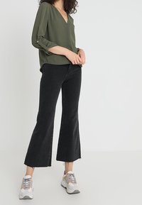 Blusa verde oliva con scollo a V e maniche arrotolate. Abbinata a jeans neri a vita alta e a campana, con sneakers con accenti grigi e pastello.