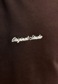 Fond en tissu brun avec texte cursif blanc "Originals Studio" brodé au centre.