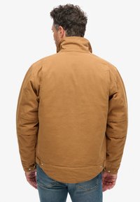 Bruine katoenen jas met een hoge kraag, gewatteerde schouders en drukknoopsluitingen aan de zoom, gecombineerd met blauwe spijkerbroek.