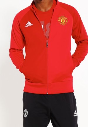 Person trägt eine rote Manchester United Zip-Jacke und schwarze Adidas-Hose mit den Händen in den Jackentaschen.