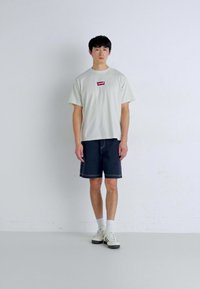 Šviesiai pilkas trumpomis rankovėmis marškinėliai su raudonu Levi's logotipu, derinami su tamsiomis džinsinėmis šortais, baltomis sportinėmis bateliais, prieš paprastą foną.