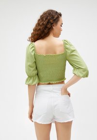 Grüner Smock-Crop-Top mit puffärmel und fransigen Kanten, kombiniert mit weißen Jeans-Shorts mit fransigem Saum.