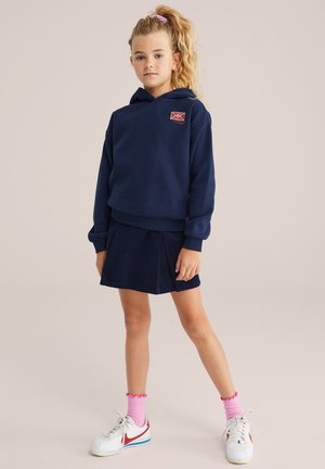 Granatowa bluza z kapturem z detalem logo, w zestawieniu z pasującą plisowaną spódnicą z korduroju. Białe sneakersy z czerwonymi akcentami i różowe skarpetki.