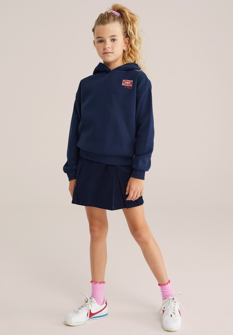 Tumši zils kapucēts džemperis ar logotipu, kombinēts ar atbilstošu samta kleitu ar pleciem. Baltas basketbola apavi ar sarkaniem akcentiem un rozā zeķēm.