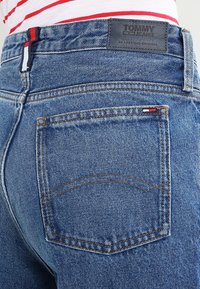 Blå denimjeans med en midjig design, med en läderlogo-patch och en distinkt röd, vit och blå detalj på bakfickan.