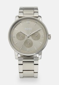 Reloj de metal con caja y banda de plata, un dial gris claro que presenta tres subdiales y marcadores numéricos en las posiciones de 12, 6 y 3.