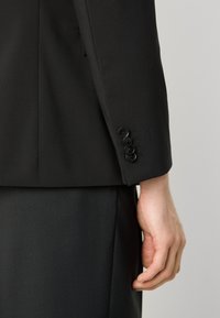 De mouw van het zwarte op maat gemaakte colbert heeft vier zwarte knopen, een gladde stofstructuur en een slim fit silhoët. Subtiele stikseldetails zijn zichtbaar.