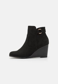 Bottine noire à talon en suède, avec un bout arrondi, une découpe sur le côté et une sangle avec un accent métallique circulaire.