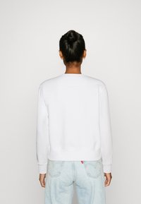 Persona con capelli scuri raccolti in un bun, che indossa piccoli orecchini a cerchio, una felpa bianca semplice e jeans azzurri chiari, in piedi contro uno sfondo bianco.