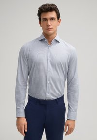 Himmelblaues Hemd mit Knöpfen, dezentem Muster, langen Ärmeln und klassischem Kragen. Kombiniert mit dunkelblauen Hosen.