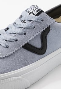 Zapatilla de ante gris con acentos negros, que presenta cordones redondos, una superficie texturizada y una suela de goma blanca. Logo visible en la lengüeta.
