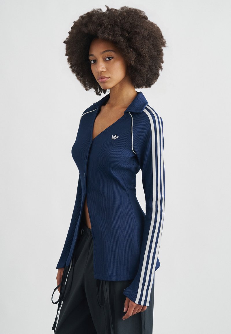 Tumši zila ribota krekla ar pogām un apkakli, ar baltām akcentu svītrām uz piedurknēm un mazu logotipu uz krūtīm.