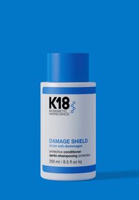 Witte plastic fles met blauwe dop en accenten. Etiket toont zwarte tekst voor "K18 DAMAGE SHIELD" en productdetails in blauw en zwart. 250 ml.