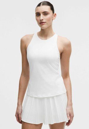 Femme portant un haut blanc sans manches et une jupe blanche plissée, posant devant un fond clair uni.