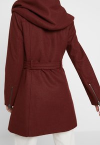 Burgundowa wełniana kurtka z dużym kołnierzem szalowym, talią w pasku oraz mankietami na zamek z gładką fakturą i dopasowanym krojem.
