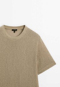 Beige lyhythihainen neulepaita, jossa on röpelökuvio ja pyöreä pääntie, makaa litteänä valkoisen taustan päällä.