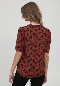 Blouse couleur rouille à manches courtes, ornée de motifs floraux noirs, avec un col froncé et un ourlet légèrement arrondi.