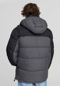 Persona con capelli ricci castani che indossa un giubbotto a blocchi di colore nero e grigio con cappuccio e jeans azzurri, in piedi con la schiena rivolta.