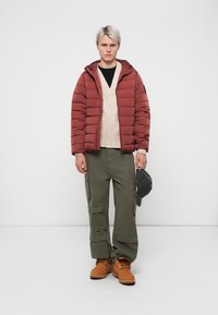 Maroon Pufferjacke mit Kapuze, darunter eine cremefarbene Strickjacke, Cargo-Hose und sandfarbene Stiefel. Halten eine graue Mütze und stehen vor einem weißen Hintergrund.