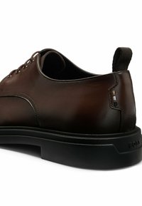 Brązowe skórzane buty wizytowe z czarną gumową podeszwą, okrągłym czubkiem, eleganckim designem i tylna pętlą do zakładania. Posiadają delikatne przeszycia i akcenty z metalowych elementów.