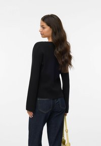 Svart stickad tröja med långa ärmar, rund halsringning och en figursydd passform, i kombination med mörka denimjeans och en gul handväska.
