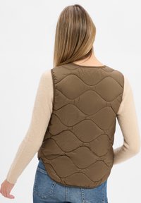 Braune gesteppte Weste mit Wellenmuster, getragen über einem beigen Wollpullover. Zeigt einen runden Halsausschnitt und eine lockere Passform.