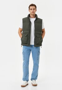 Chaleco acolchado verde oliva con cuello alto, cremallera delantera, dos grandes bolsillos, llevado sobre una camiseta blanca, combinado con pantalones cargo de un azul claro y zapatillas.