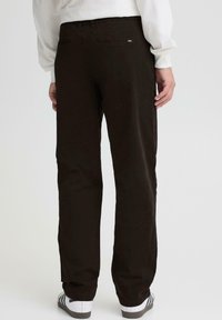 Pantalones de tela marrón duradera con un diseño de pierna recta, que cuentan con dos bolsillos traseros y una pequeña etiqueta de marca en la cinturilla, combinados con zapatos blancos.