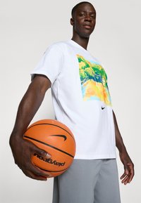 Hvid t-shirt med et farverigt grafisk print; løs pasform. Lysegrå shorts; spiller der holder en orange basketball med sorte accenter.