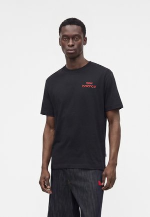 Joven con camiseta negra de New Balance con logo rojo, de pie frente a un fondo liso y claro, mirando hacia adelante con expresión neutral.