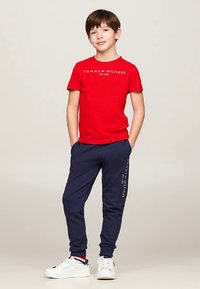 Gutt som står med hendene i lommene iført en rød Tommy Hilfiger t-skjorte, marineblå joggebukser og hvite sneakers på en ensfarget bakgrunn.